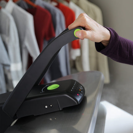 Hand using a black and green steam press on a reflective surface with clothes in the background